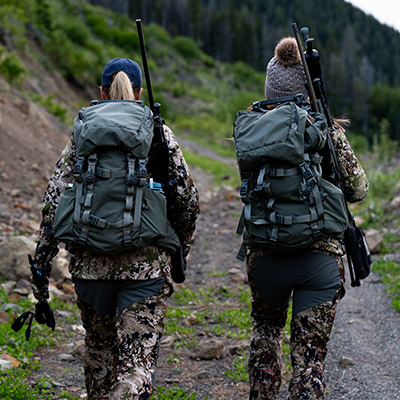 Two women hunters with packs and rifles walking down a trail.