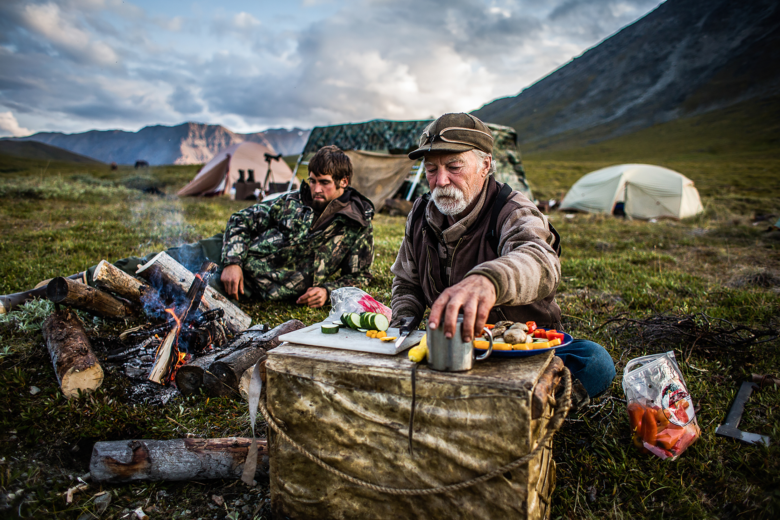 two men cooking vegetables over a camp fire